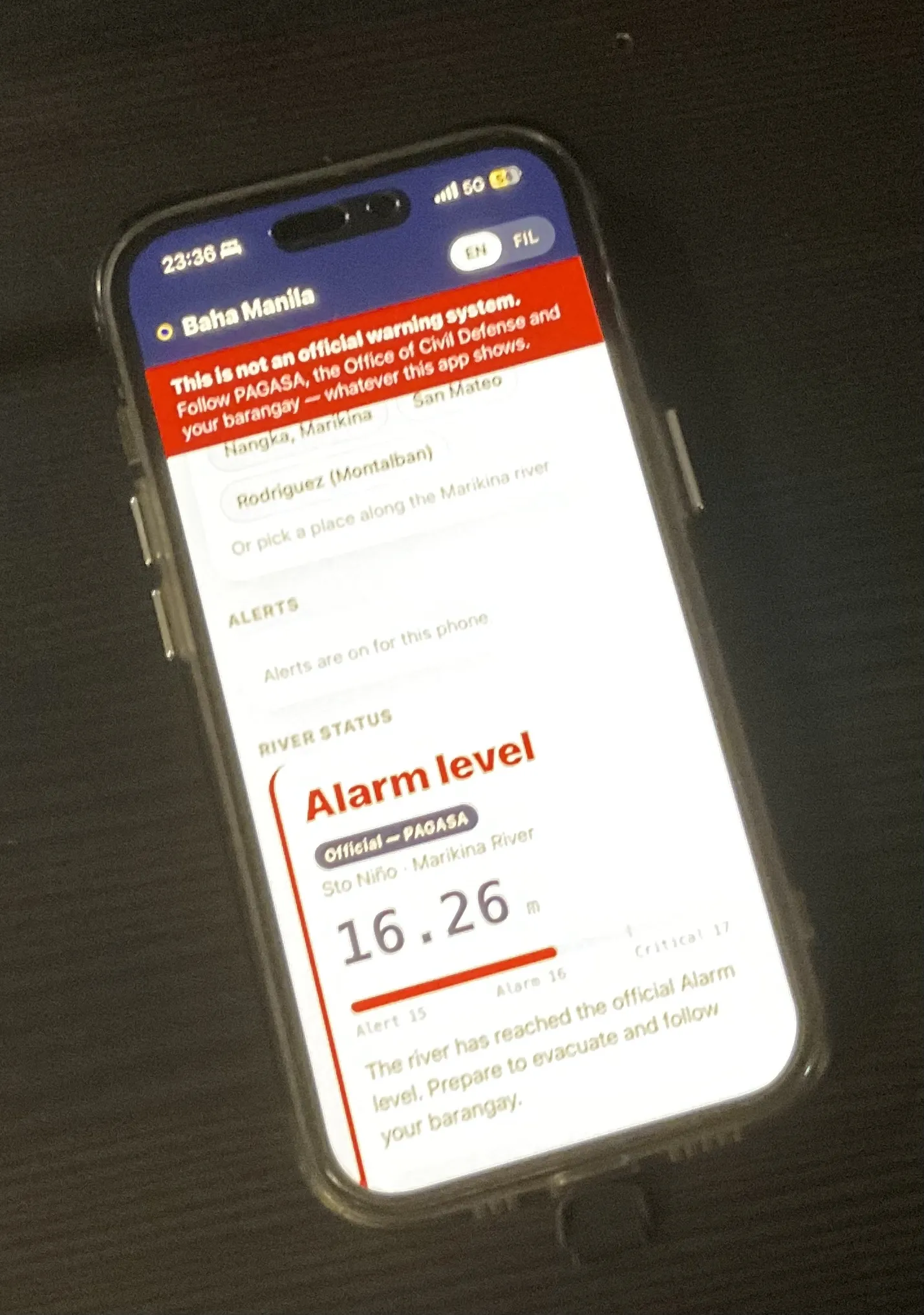 A photograph of an iPhone held over a dark desk at 23:36, showing the app in English — Baha Manila, the red not-an-official-warning-system banner, and a river status card reading "Alarm level", Official — PAGASA, Sto Niño, Marikina River, 16.26 m, with the instruction to prepare to evacuate.
